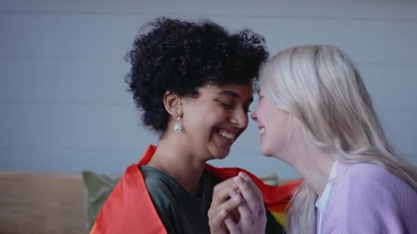 Fiatal leszbikus pár csókok kéz a kézben, ül a kanapén, visel lgbt zászló — Stock videók