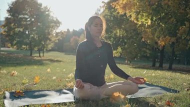 Güneşli bir sonbahar gününde şehir parkında lotus pozisyonunda oturan genç bir kadın.