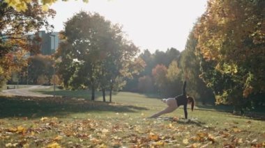 Genç bir kadın, sonbahar parkında yoga minderinin üzerinde tek bacağının üzerine tahta çakıyor.