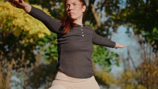 Közelkép egy fiatal nőről, aki harcos pózban áll az őszi parkban egy jóga szőnyegen. — Stock videók