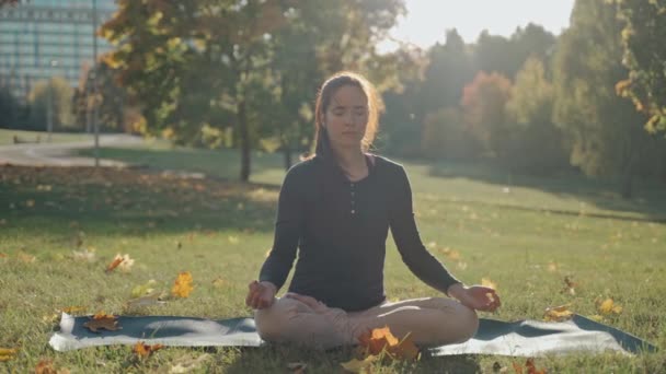 Молода жінка сидить на килимку в позі лотоса в міському парку в сонячний осінній день — стокове відео