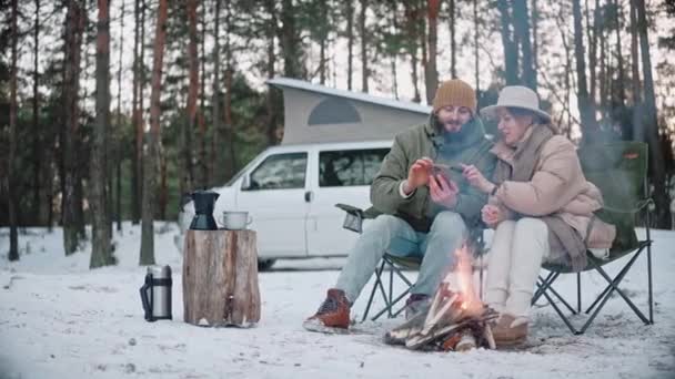Пара мандрівників сидить біля багаття, дивлячись на телефон в лісі взимку — стокове відео