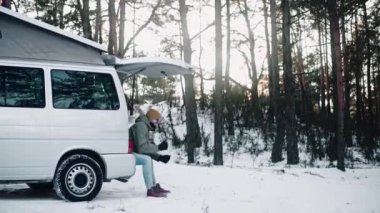 Adam kışın ağaçların arasında çatısı olan bir minibüsün arkasında oturmuş içiyor.
