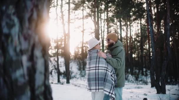 Чоловік обіймає жінку в ковдрі в лісі в зимовий сонячний день, зігріваючи її — стокове відео