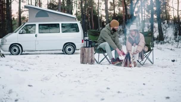 Пара сидить біля багаття в лісі взимку, насолоджуючись життям фургона — стокове відео