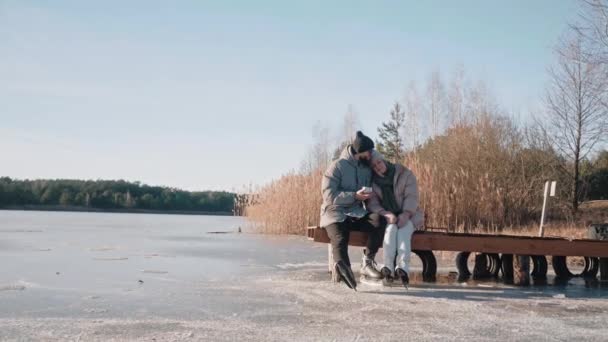 Nettes Paar sitzt im Winter auf einem Hafenbecken und trinkt heißen Tee aus der Thermoskanne und umarmt sich — Stockvideo