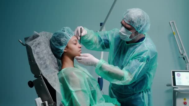 Χειρούργος αντλεί γραμμές σε ένα πρόσωπο κορίτσια πριν από ρινοπλαστική, στο μπλε γραφείο γιατρούς — Αρχείο Βίντεο