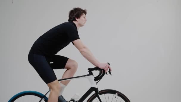 Side view of a cyclist training on an exercise bike in bright white studio — Stock Video