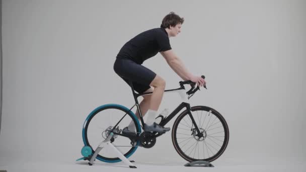 Vista lateral de um treinamento de ciclista em uma bicicleta de exercício em estúdio branco brilhante — Vídeo de Stock