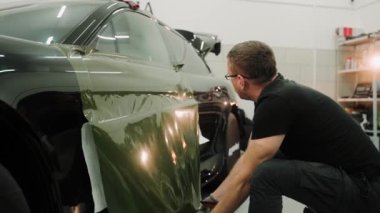 Arabanın kapısını sarmalayan, filmi indiren vinil adamın yakın çekim görüntüsü. Bir arabayı garaja sarma işlemi var. Araba ambalajı için yeşil vinil film. Yüksek kalite 4k görüntü