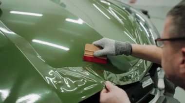 İki adam plastik kartlarla bir arabanın kaputunu koyu yeşil renge sarıyorlar. Bir araba stüdyosunda araba kaputu sarma işlemi. Parlak haki rengi yeşil araba plastiği. Yüksek kalite 4k görüntü