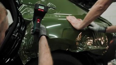 İki adam bir arabayı koyu yeşil renkte paketliyor. Bir araba stüdyosuna araba sarma işlemi. Filmi uzatmak için ısı tabancası kullanılıyor. Koyu yeşil araba plastiği. Yüksek kalite 4k görüntü