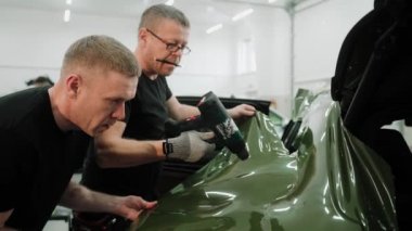 Erkekler plastikleri koyu yeşil bir arabaya sarıyorlar. Isı tabancası kullanıyorlar. Bir arabayı araba stüdyosunda ısıtma tabancası ile sarma ve filmi gerdirme işlemi. Koyu yeşil araba plastiği. Yüksek kalite 4k görüntü