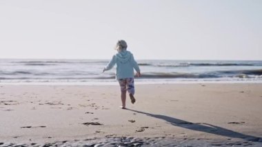 Güneşli bir günde okyanusa koşan küçük bir kız. Kumsalda serin ve güneşli bir gün. Sahilde koşan çocuğun dinamik görüntüsü. Yüksek kalite 4k görüntü