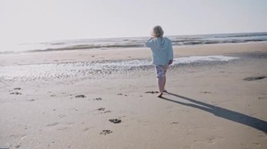 Güneşli bir günde okyanusa koşan küçük bir kız. Kumsalda serin ve güneşli bir gün. Sahilde koşan çocuğun dinamik görüntüsü. Yüksek kalite 4k görüntü