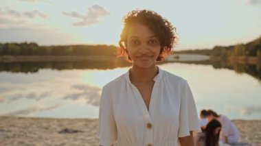 Gün batımında kumsalda kameraya bakan güzel siyah bir kız. Güzel bir genç kız göl kıyısında gün batımında eğleniyor. Kız kameraya gülümsüyor. Yüksek kalite 4k görüntü