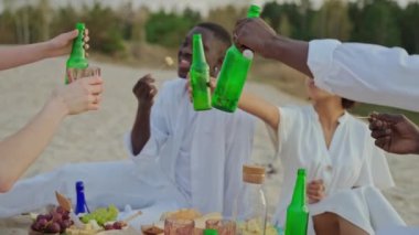 Bir grup arkadaş gün batımında plaj partisinde oturan şişelere tezahürat yapıyor. Gençler sahildeki piknikte konuşur ve içerler. Bir kadeh tost şişesi. Yüksek kalite 4k görüntü