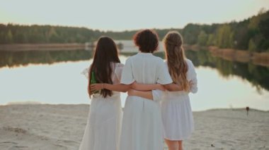 Bir grup kız arkadaş göl sahilindeki partide eğleniyorlar. Kadınlar içki içerken gülüp sarılıyorlar. İnsanlar gün batımında göl kıyısında parti veriyor. Yüksek kalite 4k görüntü