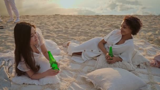 日落时 年轻姑娘们躺在海滩上聊天喝酒 美丽的年轻姑娘们在湖滨的夕阳西下玩得很开心 日落时的海滩派对高质量的4K镜头 — 图库视频影像