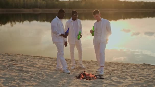 一群在湖边玩乐的男性朋友在篝火边喝酒 三个人在日落时在湖边举行聚会 穿着白衣的人在聚会 高质量的4K镜头 — 图库视频影像