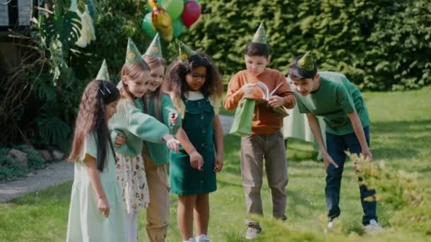 在阳光灿烂的一天 孩子们在后院的生日派对上玩得很开心 绿色主题的生日派对 男孩和女孩们戴着生日帽看着草坪上的什么东西 高质量的4K镜头 — 图库视频影像