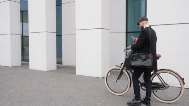Takım elbiseli yakışıklı bir adam iş merkezinin yakınında telefonla konuşuyor. Şapka ve omuz çantası takan bir adam, siyah eski moda bir bisiklet tutuyor. Yüksek kalite 4k görüntü