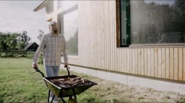 Adam güneşli bir günde arka bahçede el arabasında ahşap bir ağaç kabuğu sürüyor. Gömlek ve şapka giyen bir adam. Arka bahçede çalışan İskandinav yaşam tarzı adamı. Yüksek kalite 4k görüntü