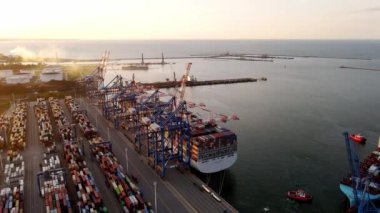 Aerial view of an industrial port with cargo ship with containers at sunset. Sun setting at the sea. Camera flying over port with shipping containers. High quality 4k footage