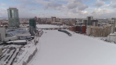 Modern yüksek binaları ve donmuş nehri olan büyük bir Rus şehrinin havadan görünüşü. Kamera nehrin üzerindeki yol köprüsüne zumluyor. Köprü yeniden inşa ediliyor. Bulutlu gün.