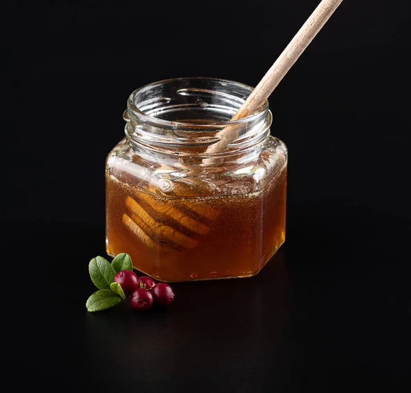 Beautiful delicious transparent honey flows down from the honey spoon into a glass jar. A branch of fresh young cranberries. On a black background with reflection