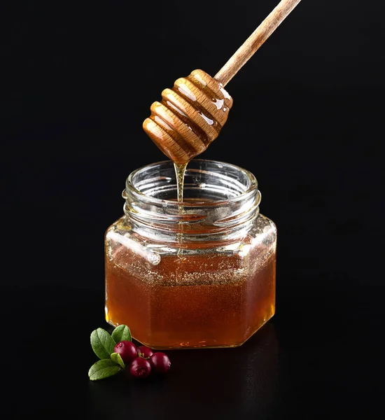 Beautiful delicious transparent honey flows down from the honey spoon into a glass jar. A branch of fresh young cranberries. On a black background with reflection