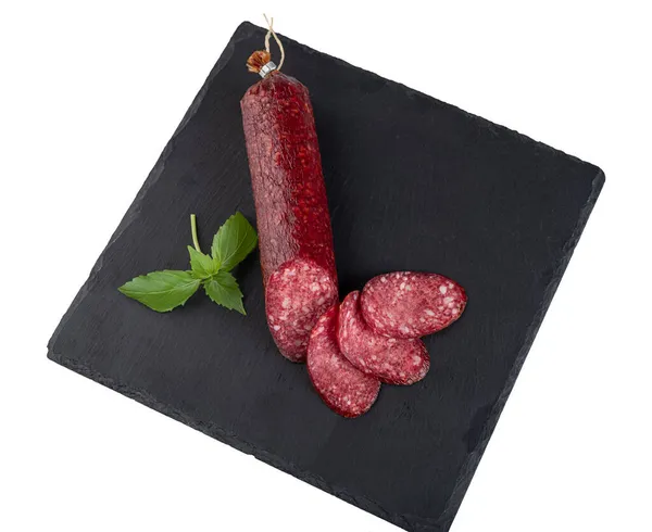 Slices of smoked sausage and a sausage stick with fresh basil on a black slate cutting board, isolated on a white background