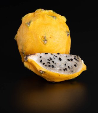 Tam ve doğranmış sarı, ballı ejderha meyvesi parlak siyah bir arka planda yansımalı.