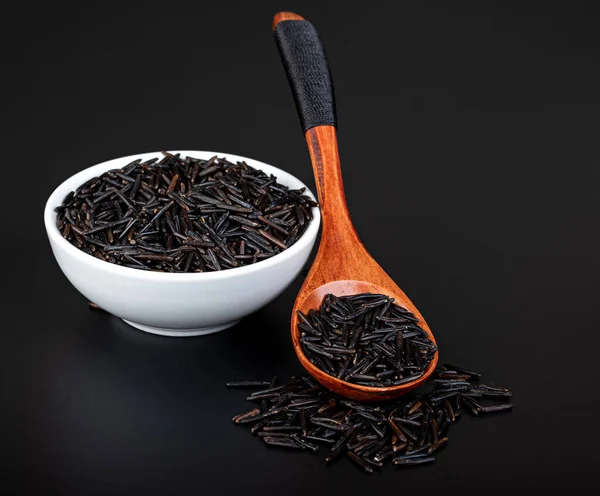 Siyah arka planda tahta kaşıkla beyaz bir kasede siyah pirinç..