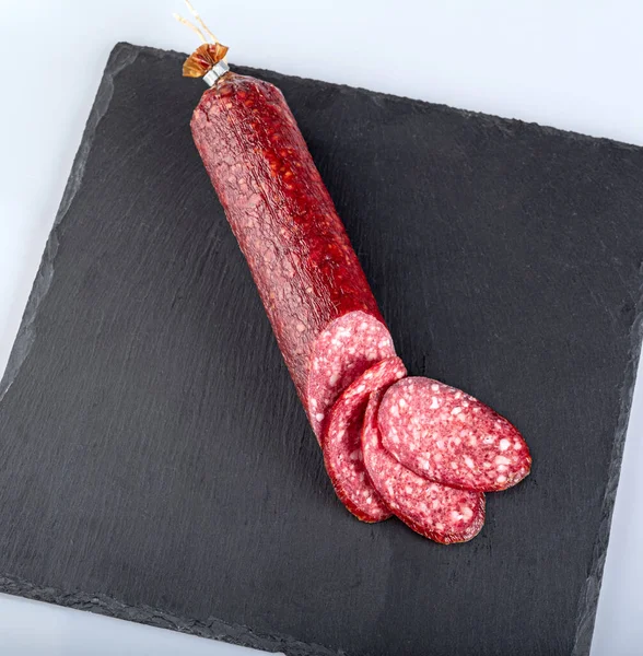 Slices of smoked sausage and a sausage stick on a black cutting board, isolated on a white background
