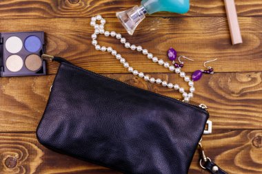 Ahşap arka planda kadın aksesuarları. Debriyaj çantası, parfüm şişesi, göz farı, inci kolye ve tahta masada küpeler. Güzellik ve moda kompozisyonu. Üst manzara, düz uzanma