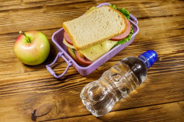 Sandviçli beslenme çantası, bir şişe su ve ahşap masada elma.