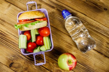 Bir şişe su, elma ve beslenme çantası, içinde hamburgerler ve ahşap bir masada taze sebzeler. Üst görünüm