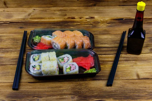 Set of sushi rolls in plastic boxes, soy sauce and chopsticks on wooden table. Sushi for take away or delivery of sushi in plastic containers