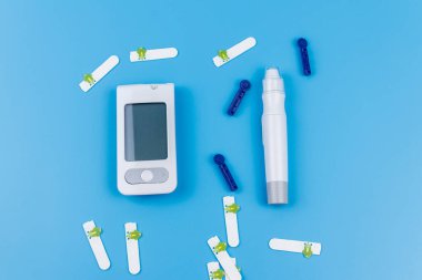 Dijital glukometre, tükenmez kalem, tek kullanımlık iğneler ve pastel mavi arka planda test şeritleri. Üst Manzara. Diyabet kavramı