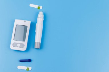 Dijital glukometre, tükenmez kalem, tek kullanımlık iğneler ve pastel mavi arka planda test şeritleri. Üst görünüm, boşluğu kopyala. Diyabet kavramı