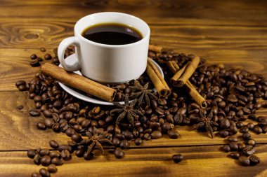 Bir fincan kahve, kavrulmuş kahve çekirdekleri, yıldız anason ve ahşap masada tarçın çubukları.