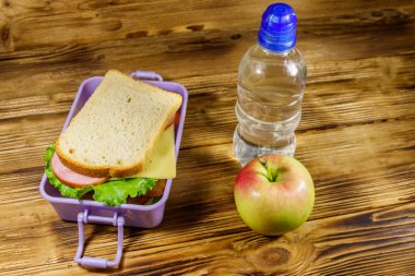 Sandviçli beslenme çantası, bir şişe su ve ahşap masada elma.