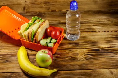 Bir şişe su, elma, muz ve sandviç ve taze sebzelerle birlikte ahşap bir masada.