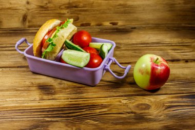 Elma, beslenme çantası, hamburgerler ve ahşap bir masada taze sebzeler.