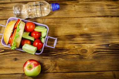 Bir şişe su, elma ve beslenme çantası, içinde hamburgerler ve ahşap bir masada taze sebzeler. Üst görünüm, alanı kopyala