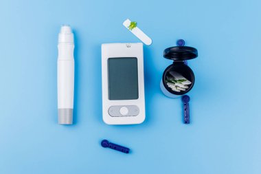 Dijital glukometre, tükenmez kalem, tek kullanımlık iğneler ve pastel mavi arka planda test şeritleri. Üst Manzara. Diyabet kavramı