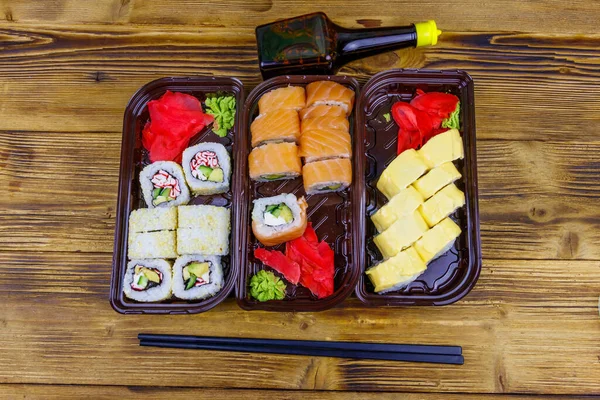 Set of sushi rolls in plastic boxes, soy sauce and chopsticks on wooden table. Top view. Sushi for take away or delivery of sushi in plastic containers