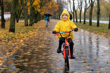 Sarı yağmurluklu çocuk sonbahar parkında bisiklet sürüyor. Yağmurda bisiklet süren çocuk. Anaokulunun mutlu portresi.