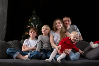 Genç ebeveynler ve üç küçük çocuk Noel arifesinde Noel ağacının yanındaki koltukta, karanlık oturma odasında oturuyorlar.